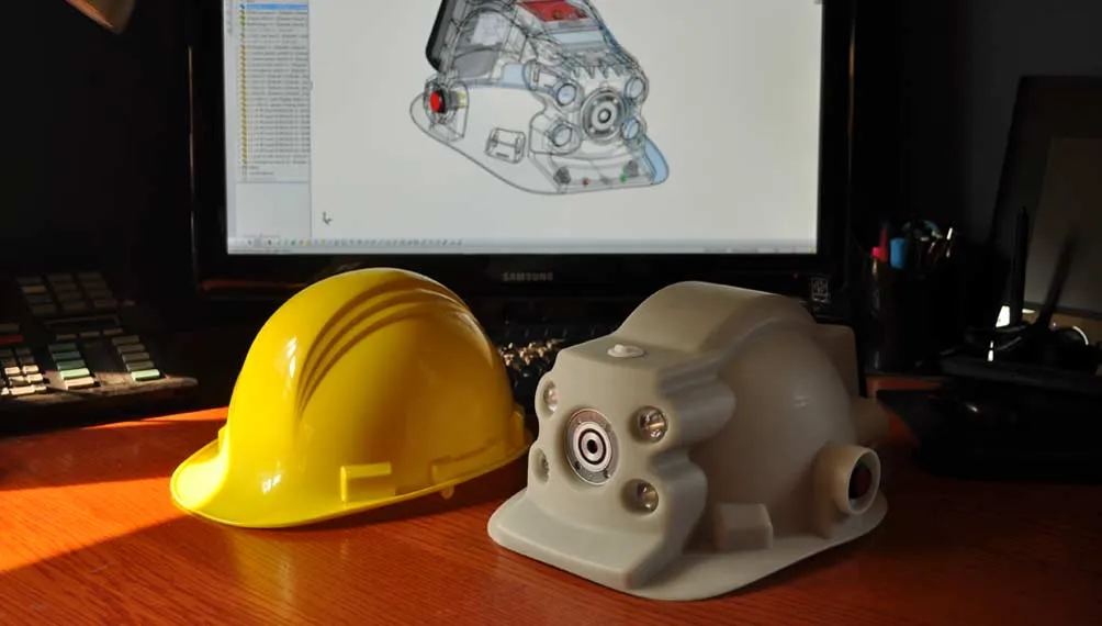 Yellow hard hat and SafeXtract helmet prototype on a desk in front of a CAD model on screen